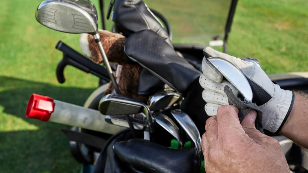 Hands in golf gloves expertly demonstrating how to clean golf clubs, using a cloth on an iron, while a set of pristine golf clubs with covers sits in a bag on the lush course, showcasing the importance of a good golf club cleaner.